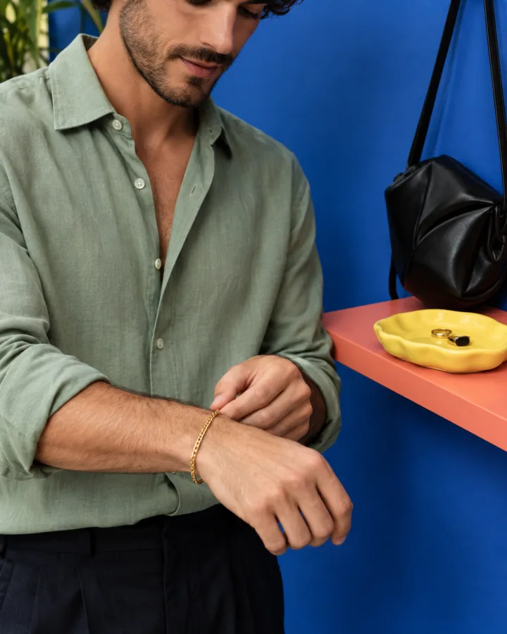 A man fastening a gold bracelet beside a black bag against a cobalt wall.